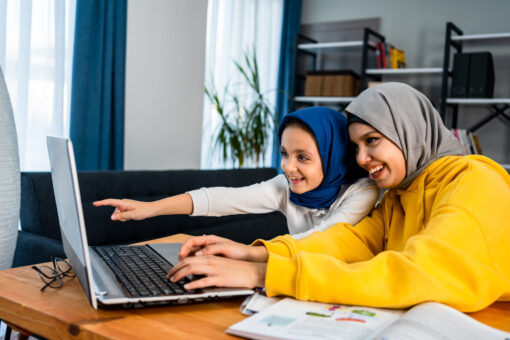 A woman and small girl sit at a desk looking at a laptop. They are both smiling.