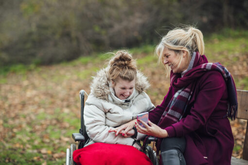 A woman sitting down shows something on her mobile phone to another woman, who is sitting in a wheelchair. They are both wrapped up warm and smiling in the park.